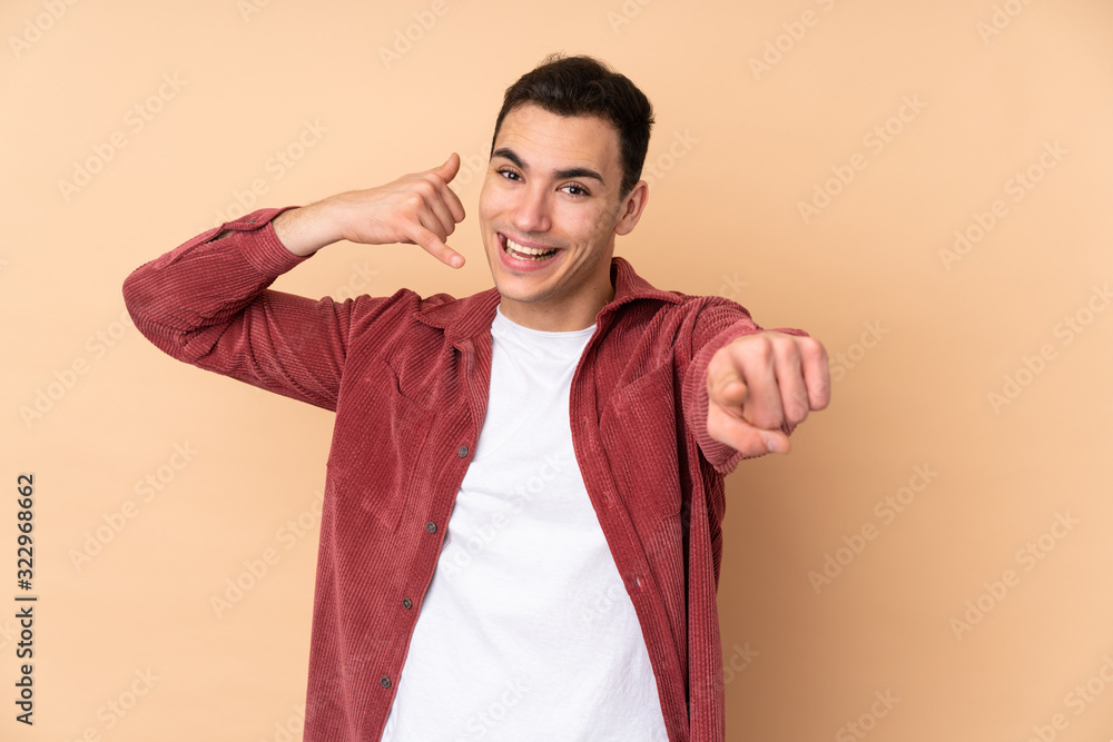 Young caucasian handsome man isolated on beige background making phone gesture and pointing front Young caucasian handsome man isolated on beige background making phone gesture and pointing front
