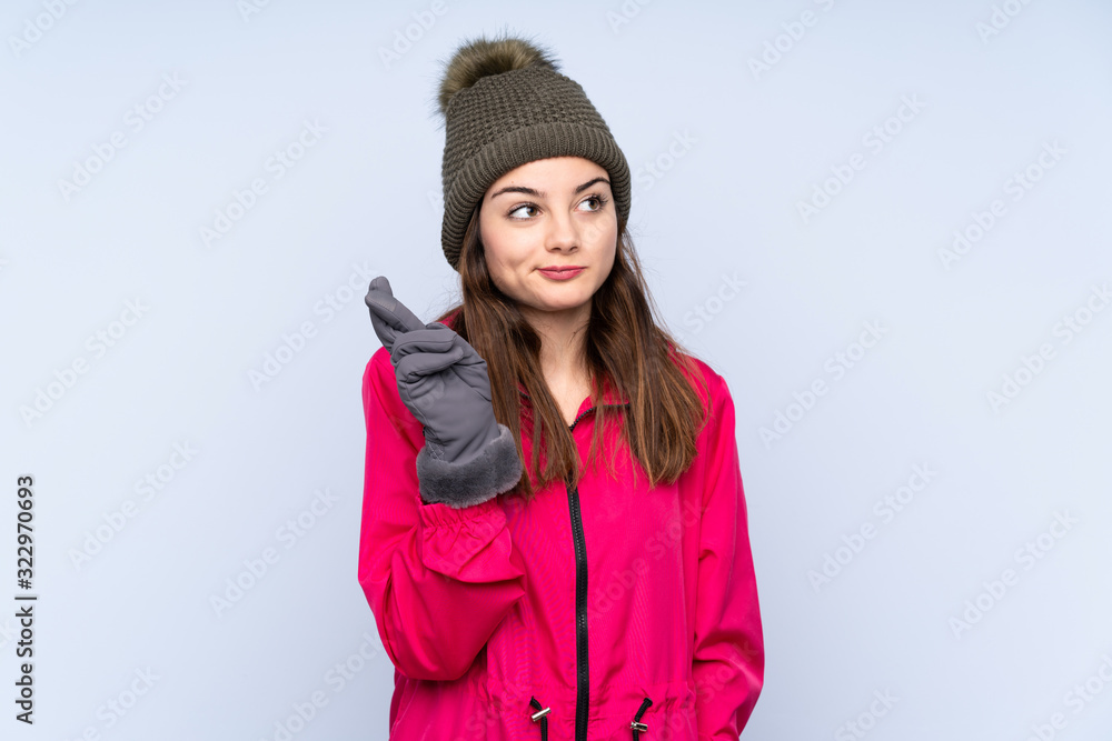 Young girl with winter hat isolated on blue background with fingers crossing and wishing the best Young girl with winter hat isolated on blue background with fingers crossing and wishing the best