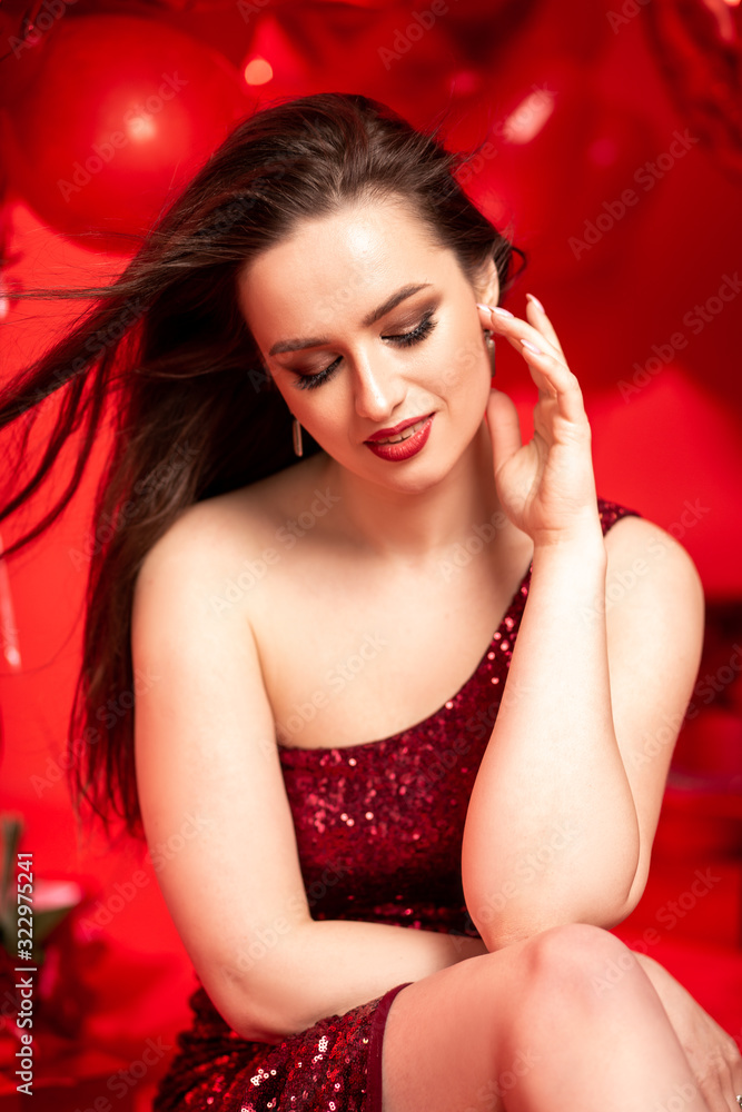 Beautiful young woman in red evening dress posing over red background with big heart shape balloons. Happy Valentine's Day concept
