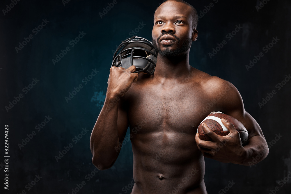 portrait photo of dark-skinned young man with nude torso on a dark background he holds a rugby helmet on his shoulder and rugby ball in his arm he looks up