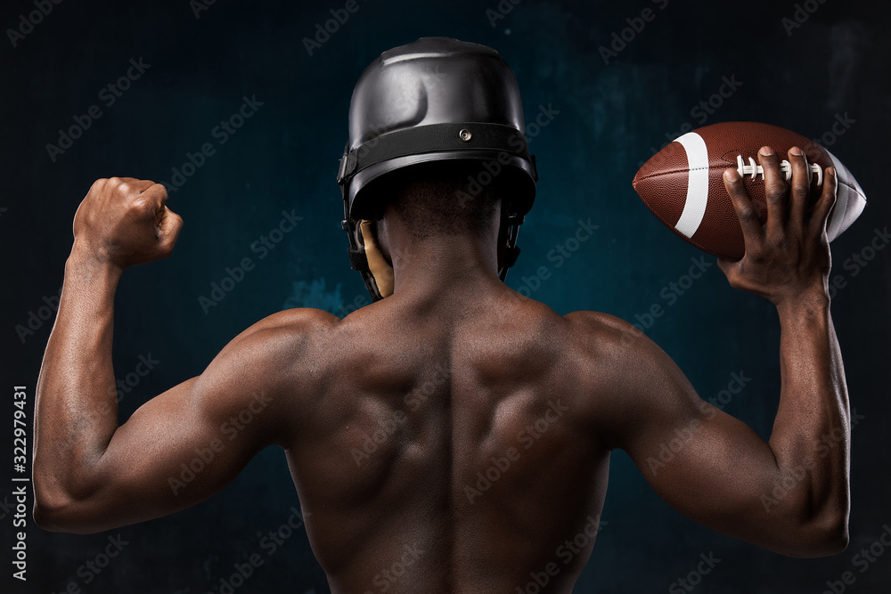 portrait photo from the back of dark-skinned young man with nude torso on a dark background he has a rugby helmet on head and holds a rugby ball up in his arm