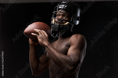 portrait photo of dark-skinned young man with nude torso on a dark background he has a rugby helmet on head and holds a rugby ball up in his arm © monchak