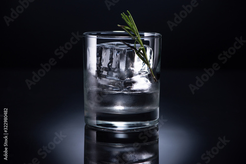 transparent glass with ice cube, rosemary and vodka on black background
