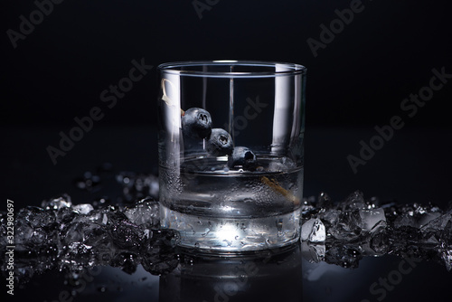 transparent glass with vodka and blueberries near smashed ice on black background