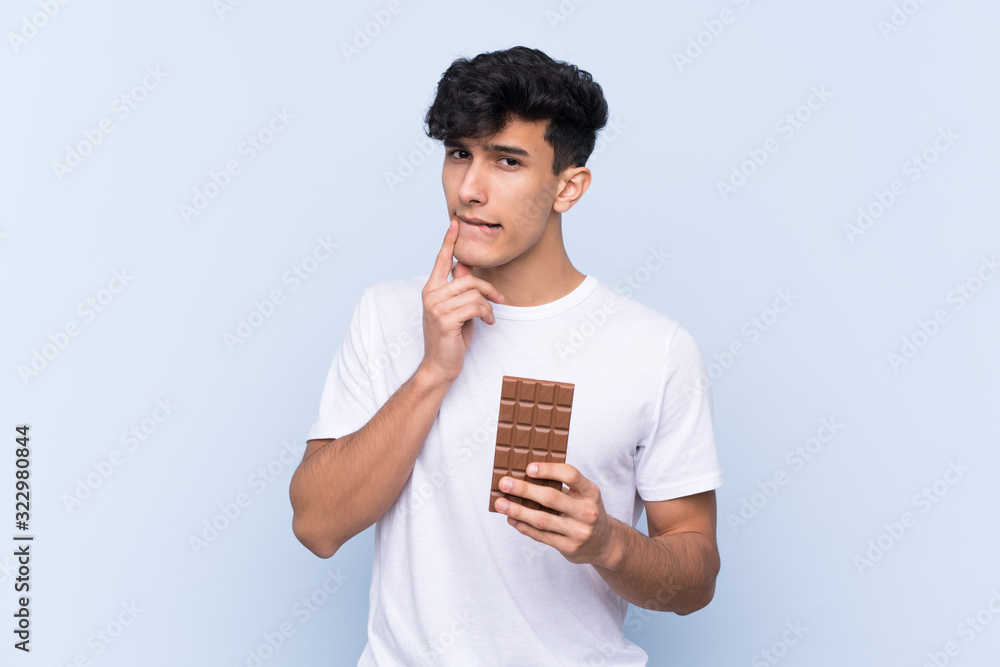 Young Argentinian man over isolated blue background taking a chocolate tablet and having doubts Young Argentinian man over isolated blue background taking a chocolate tablet and having doubts