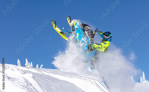 elite sports snowmobiler rides and jumps on steep mountain slope with swirls of snow storm. background of blue sky leaving a trail of splashes of white snow. bright snowmobile and suit without brands