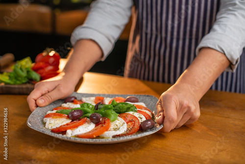 Female chef is placing a plate of fresh, delicious mozzarella and tomato salad on a wooden table.