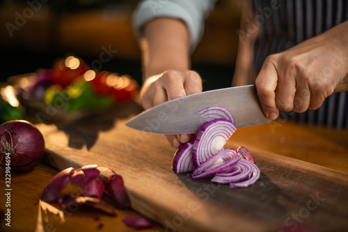 Female chef is precisely slicing red onions on a wooden cutting board in a restaurant. 