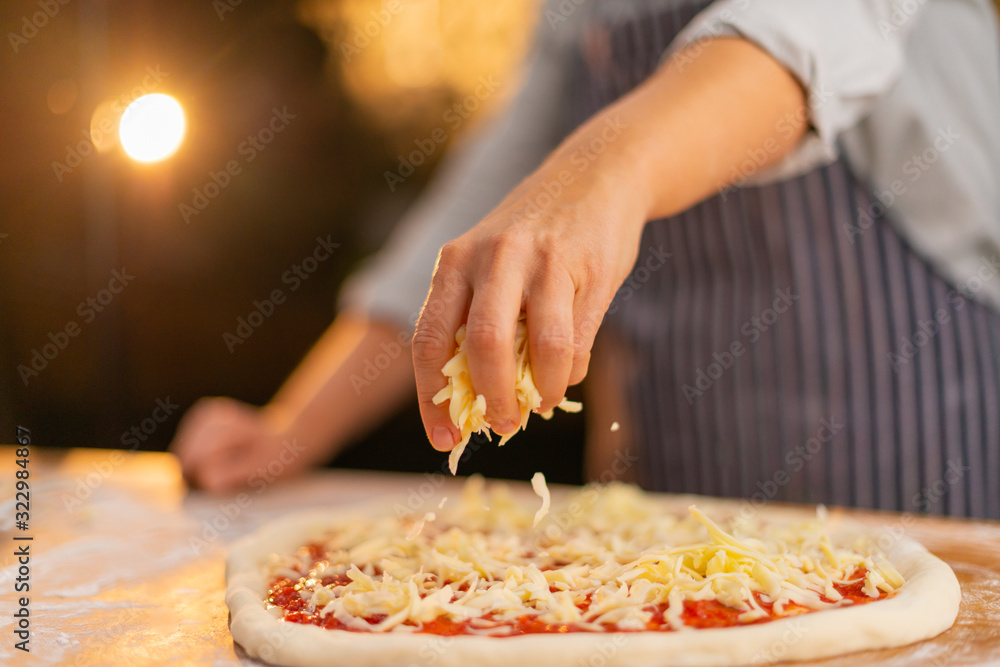 Female chef is preparing a traditional italian pizza and she is pouring cheese on the dough.