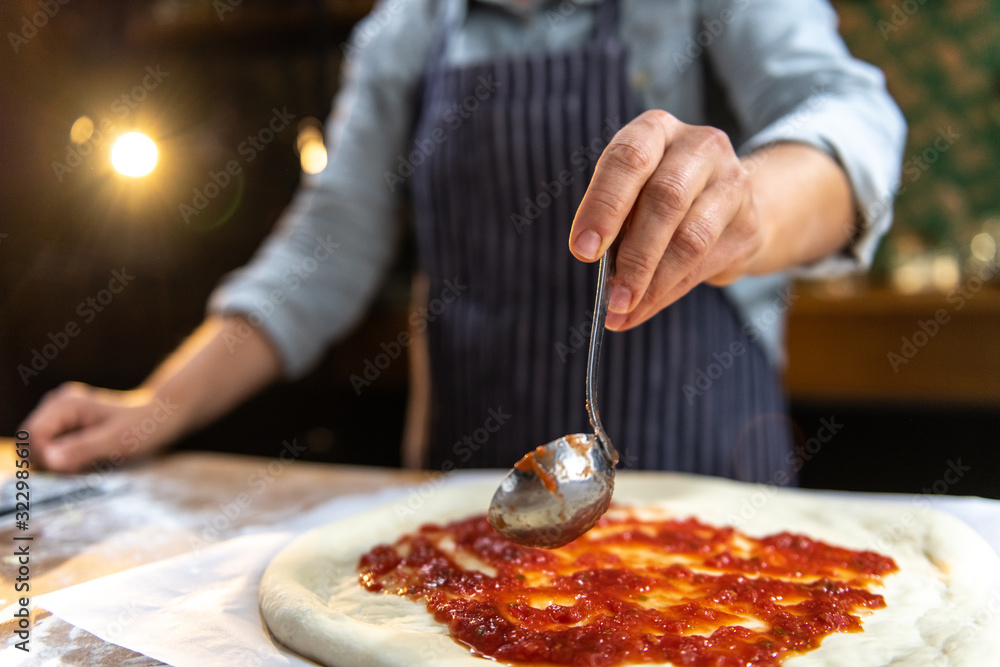 Female chef is pouring freshly made tomato sauce over a traditional, homemade pizza.
