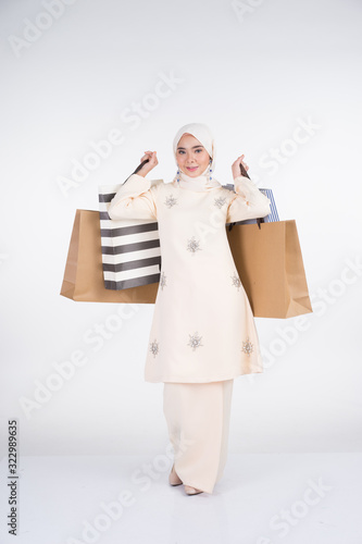 A beautiful Muslim female model in a Asian traditional dress kurung Pahang carrying shopping bags isolated on white background. Eidul fitri festive preparation shopping concept. Full length portrait.