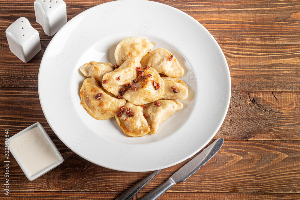 Dumplings and ravioli stuffed with meat. Sprinkle sliced smoked tomatoes. Served on a large white plate. Next to a bowl of sour cream. On a wooden background. Dumplings and ravioli stuffed with meat. Sprinkle sliced smoked tomatoes. Served on a large white plate. Next to a bowl of sour cream. On a wooden background.