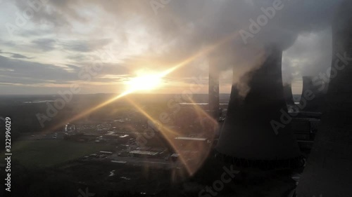 Aerial view of power station cooling towers dolly right across burning smoke emissions with atmospheric glowing sunrise.