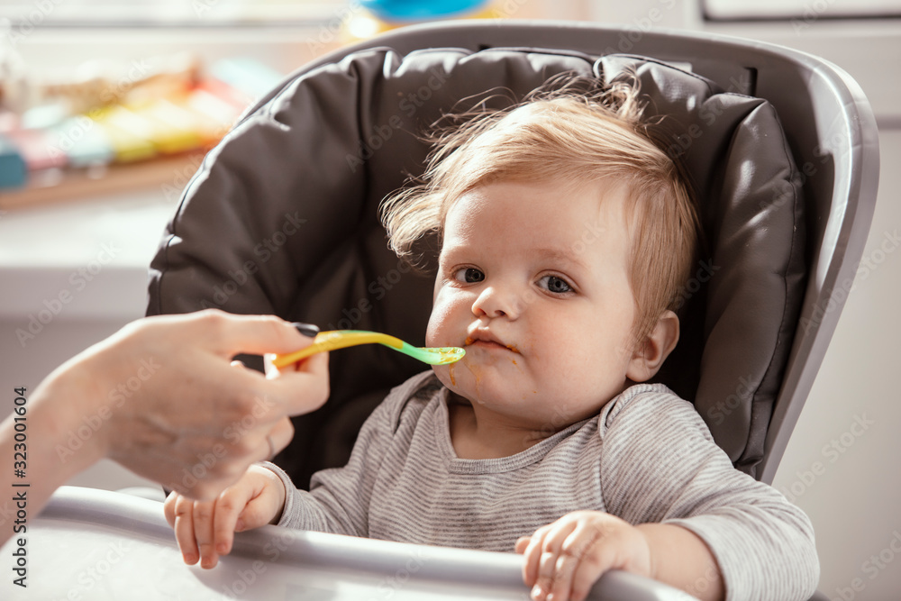 Beautiful baby in the child dining chair eat healthy food. Mother's hands. Child eat vegetable puree. Lifestyle. Sunny day. Beautiful baby in the child dining chair eat healthy food. Mother's hands. Child eat vegetable puree. Lifestyle. Sunny day.