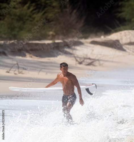 Young handsome man wet all over body,walking on the beach,holding serf board righe side,show fit and firm muscle,reflection of sunlight shining on his skin