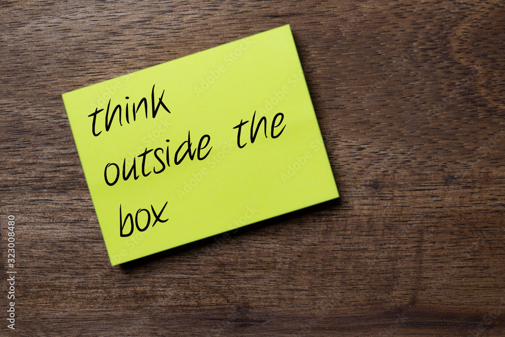 yellow self-adhesive note sticker diagonally on a dark wooden table top with stylized handwriting with the slogan think out of the box yellow self-adhesive note sticker diagonally on a dark wooden table top with stylized handwriting with the slogan think out of the box
