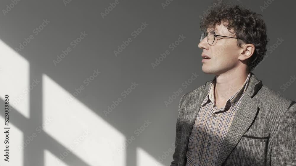 Man with glasses, with curly hair, in gray jacket, in white room with shadow from window on the wall, walks from side to side, holds pencil to his lips and reflects on another project or business idea Man with glasses, with curly hair, in gray jacket, in white room with shadow from window on the wall, walks from side to side, holds pencil to his lips and reflects on another project or business idea