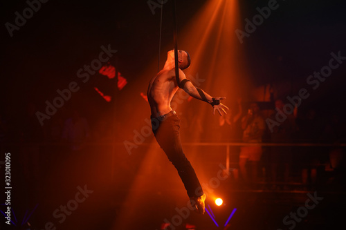 Sports man with a naked torso balances on straps backlit in red lights, circus show © Dima