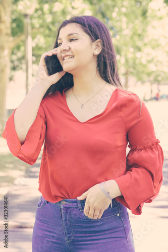 Smiling pretty young woman talking on mobile phone in park. Beautiful lady wearing red blouse and standing with green lawn and trees in background. Communication and nature concept. Front view.