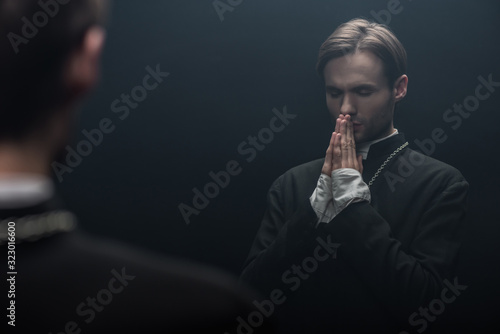 Фотография young serious catholic priest praying with closed eyes near own reflection isola