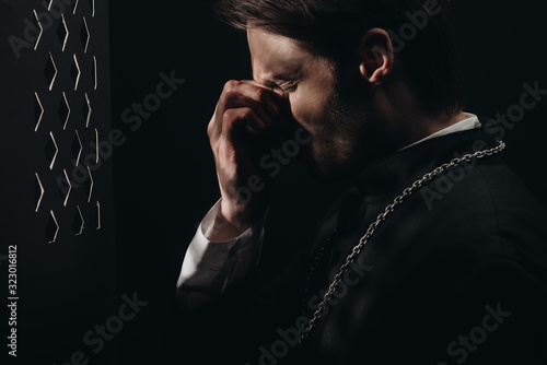 Fotografija young tense catholic priest touching face with closed eyes near confessional gri