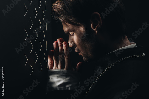 Fotótapéta young catholic priest praying with closed eyes near confessional grille in dark