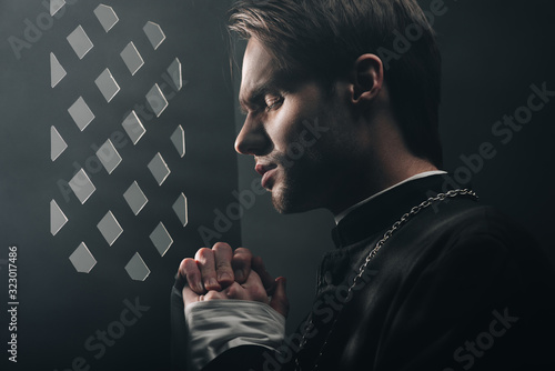 young thoughtful catholic priest praying with closed eyes in dark near confessional grille with rays of light photo