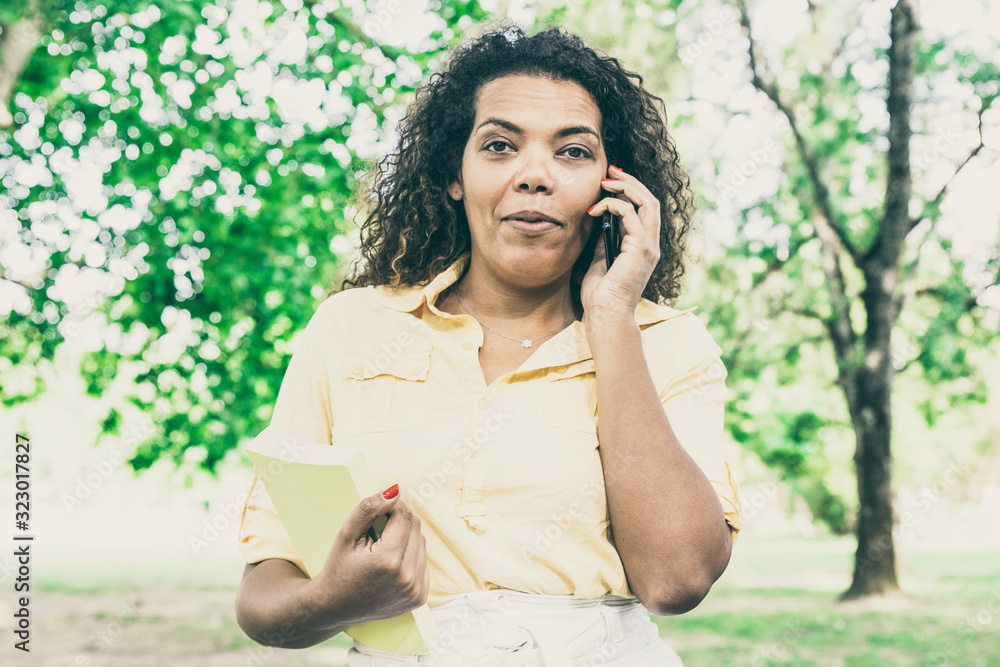Serious woman talking on phone and holding book in city park. Young woman standing with blurred trees and lawn in background. Summer and communication concept. Front view. Serious woman talking on phone and holding book in city park. Young woman standing with blurred trees and lawn in background. Summer and communication concept. Front view.