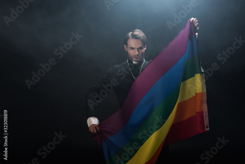 Картината върху платно confident catholic priest holding lgbt flag while looking at camera on black bac