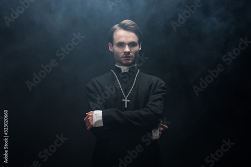 young serious catholic priest standing with crossed arms on black background with smoke photo