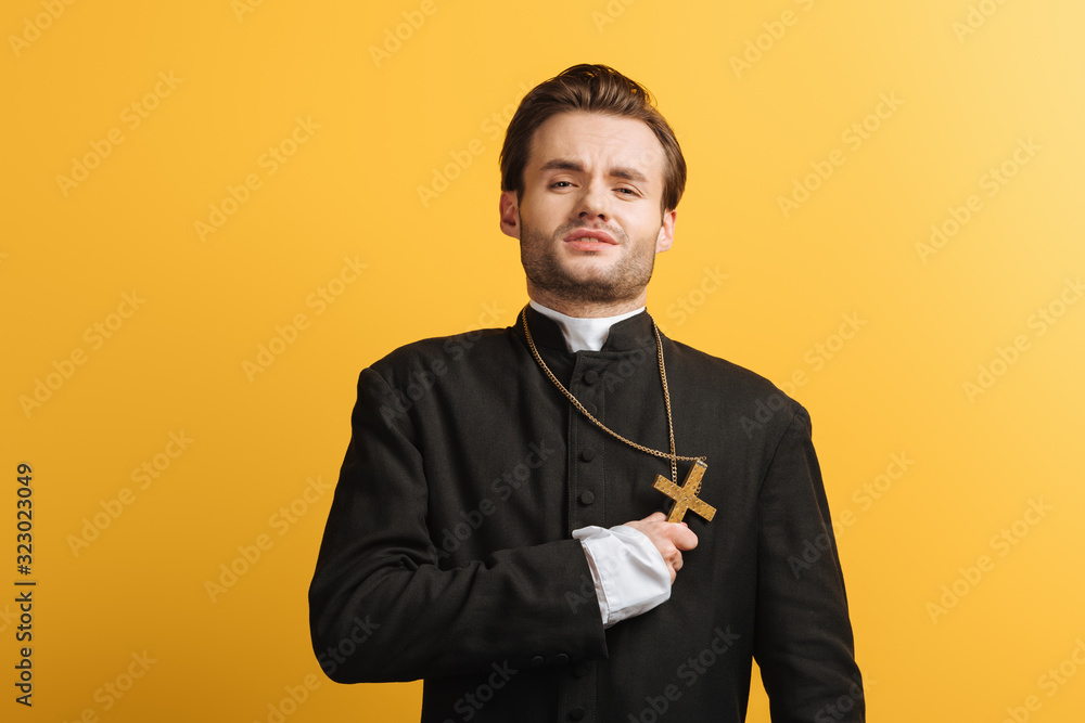 young catholic priest with exhausted face expression holding holy christ near heart isolated on yellow young catholic priest with exhausted face expression holding holy christ near heart isolated on yellow