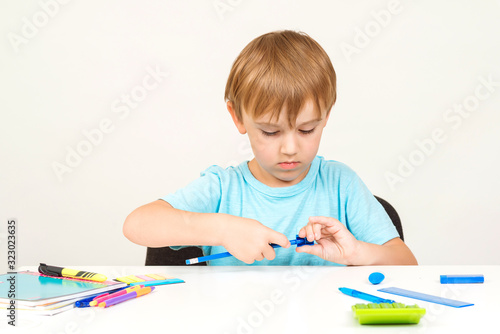 Schoolboy is sharpening the pencil. Pupil sitting at white desk with school supplies. Kid doing homework