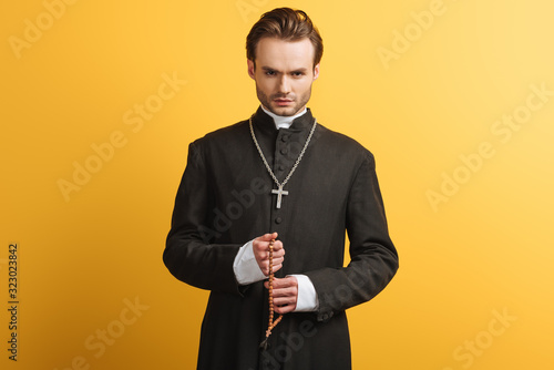 Εκτύπωση καμβά young confident catholic priest holding wooden rosary beads isolated on yellow