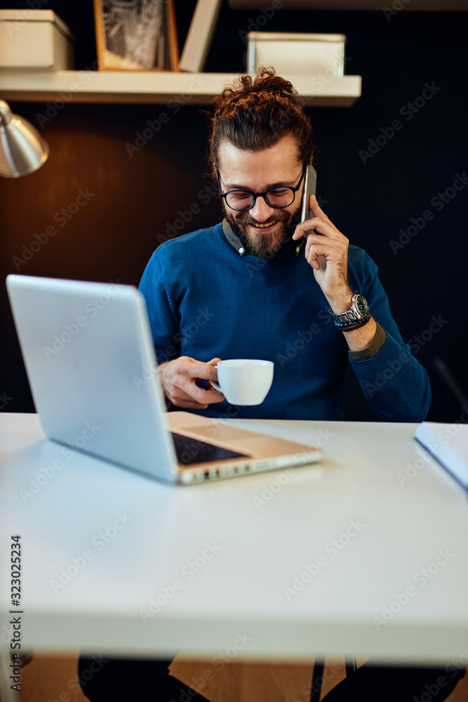 Young laughing caucasian bearded blogger sitting in his office, talking on the phone, holding cup of coffee and taking a break from work. Young laughing caucasian bearded blogger sitting in his office, talking on the phone, holding cup of coffee and taking a break from work.