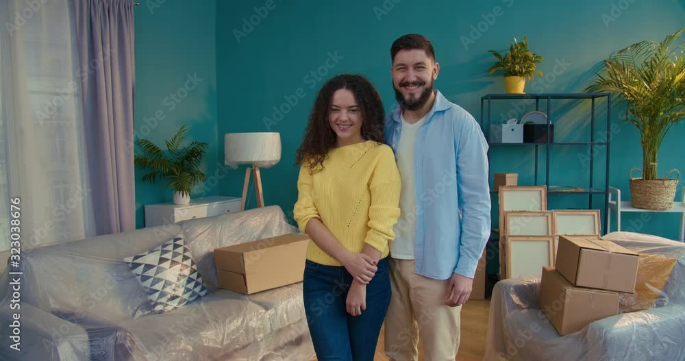 Portrait of good looking happy couple in living room with boxes looking at each other smiling to camera. Boyfriend and girlfiend moving in together in new apartment. Removing concept. Portrait of good looking happy couple in living room with boxes looking at each other smiling to camera. Boyfriend and girlfiend moving in together in new apartment. Removing concept.
