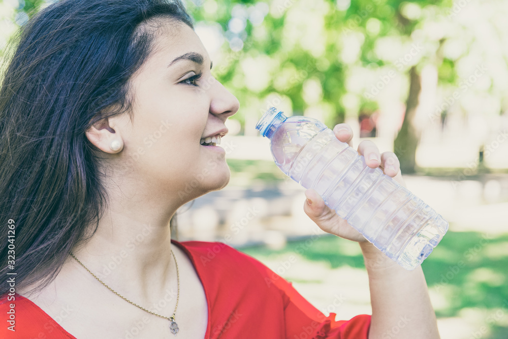 Smiling pretty young woman drinking water from bottle in park. Beautiful lady wearing red blouse with blurred green lawn and trees in background. Thirst and nature concept. Side view. Smiling pretty young woman drinking water from bottle in park. Beautiful lady wearing red blouse with blurred green lawn and trees in background. Thirst and nature concept. Side view.