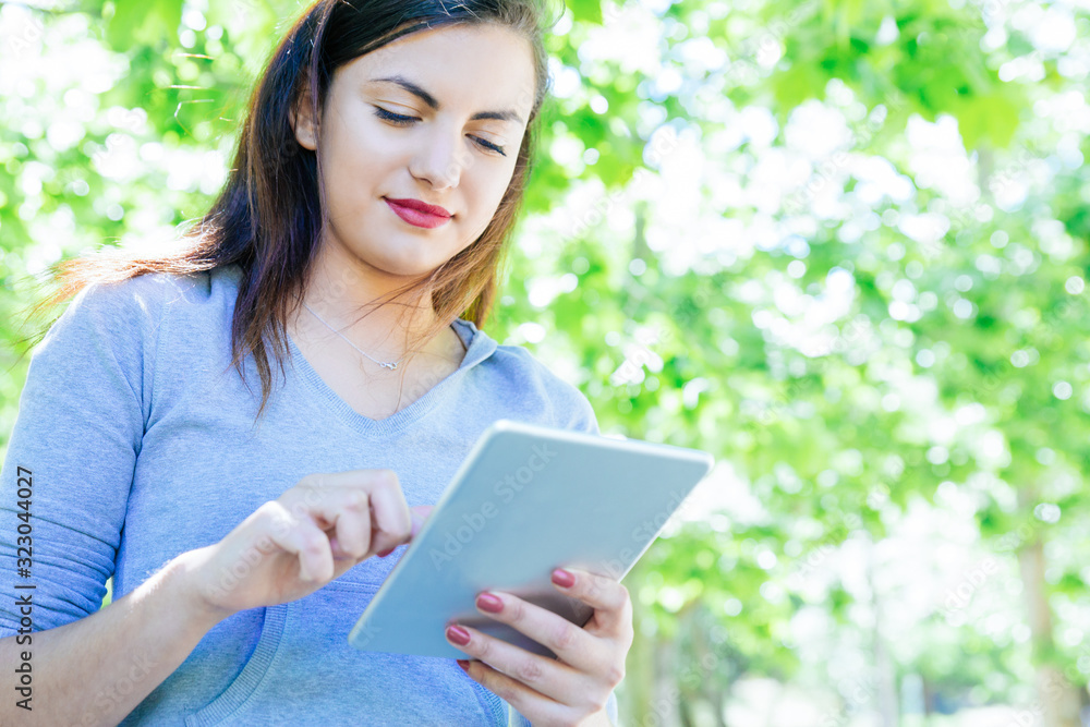 Content pretty young lady using tablet computer in park. Beautiful woman wearing hoodie and standing with green trees in background. Technology and nature concept. Front view. Content pretty young lady using tablet computer in park. Beautiful woman wearing hoodie and standing with green trees in background. Technology and nature concept. Front view.