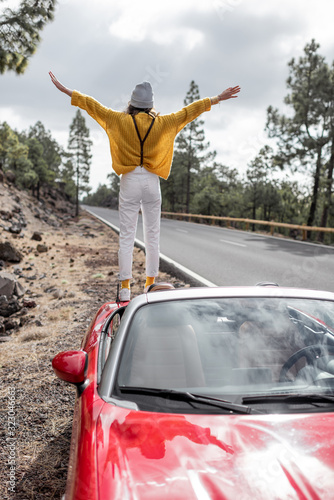 Young stylish woman traveling by car, enjoying beautiful landscapes while standing on the roadside, backside view. Carefree lifestyle and traveling concept