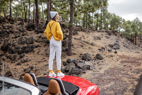Young stylish woman traveling by car, enjoying beautiful landscapes while standing on the roadside in the forest. Carefree lifestyle and traveling concept