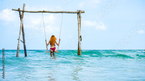 Beach summer vacation swing on th sea with young beautiful girl sitting on beach swing blue sea.Red haired woman swinging on the beach on Phu Quoc island, Vietnam. Happy on tropical ocean swing.