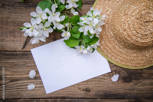 A branch of a blooming apple tree with white flowers, a sheet of paper and a straw hat on a wooden background, with a copy space