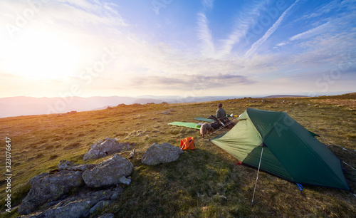 A hiker rests outside their tent while camping on a mountain summit at sunset. Rampsgill Head in the Lake District.