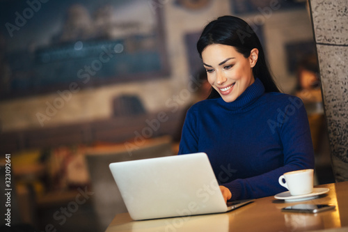 Attractive young cheerful woman having a cup of coffee and working with laptop in cafe