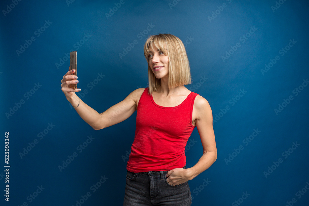 Fototapeta premium Beauty fashion portrait of slim blonde woman in red shirt on a blue background in studio making a selfie with smartphone. Social networks, selfie addiction