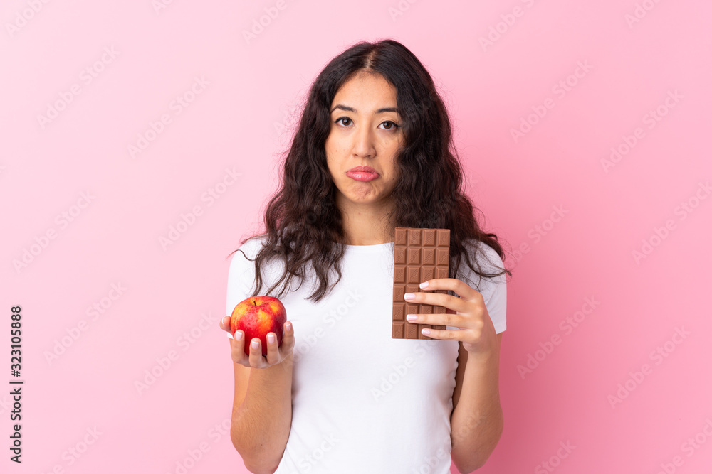 Spanish Chinese woman over isolated pink background taking a chocolate tablet in one hand and an apple in the other Spanish Chinese woman over isolated pink background taking a chocolate tablet in one hand and an apple in the other