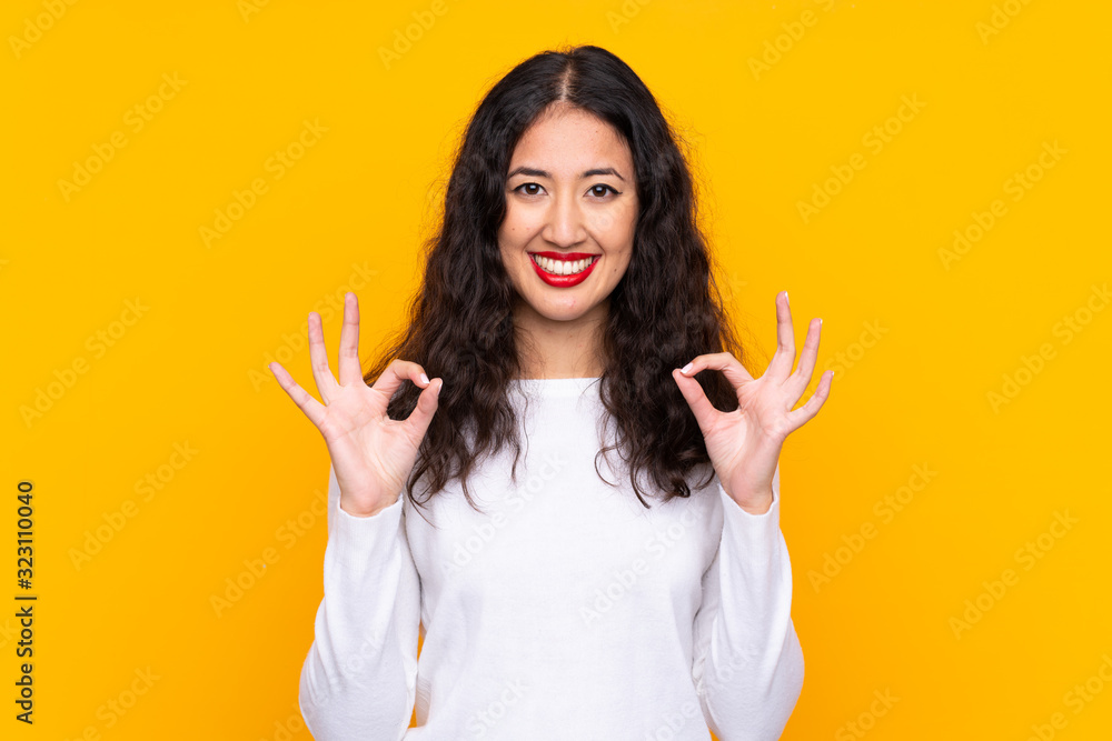 Mixed race woman over isolated yellow background showing an ok sign with fingers Mixed race woman over isolated yellow background showing an ok sign with fingers