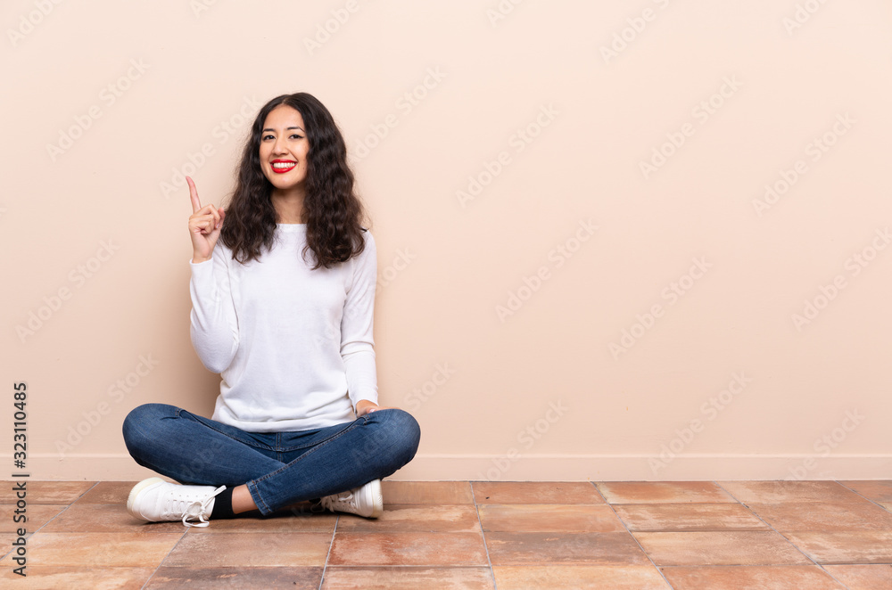 Young woman sitting on the floor showing and lifting a finger in sign of the best Young woman sitting on the floor showing and lifting a finger in sign of the best