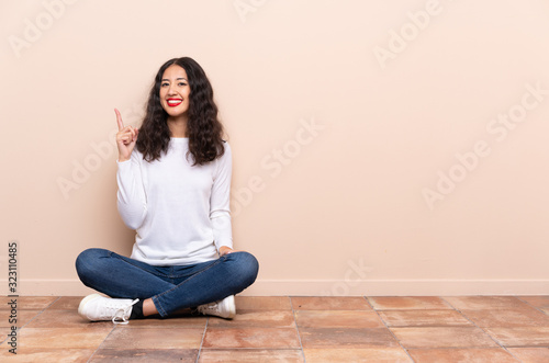 Young woman sitting on the floor showing and lifting a finger in sign of the best © luismolinero