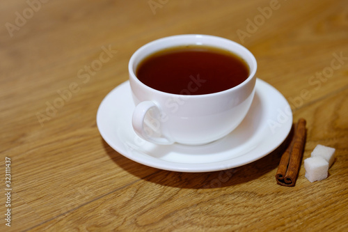 Classic white cup of black tea with saucer, teapot, sugar and cinnamon on the wooden table background. Breakfast drink at home or cafe.