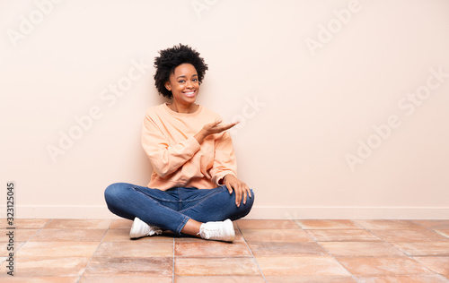 African american woman sitting on the floor presenting an idea while looking smiling towards © luismolinero